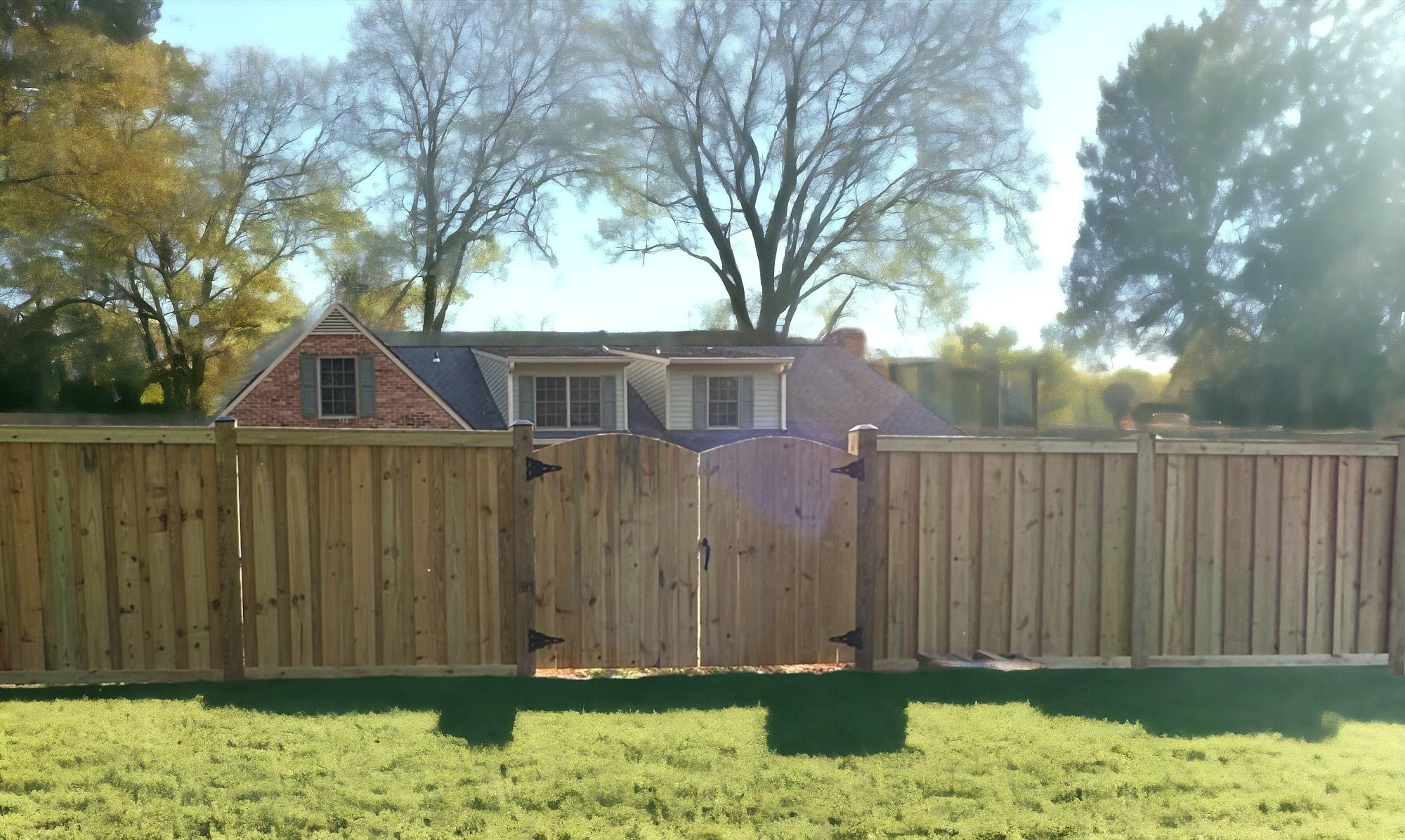 New wooden privacy fence with arched double gate opening in a residential backyard, with construction materials scattered on the ground, houses and trees in the background, and sunlight peeking through the foliage.