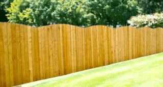 A warm-toned wooden fence with a scalloped top design stands in front of a lush garden. The fence's curved peaks create a rhythmic pattern across the image, and it is set against a background of vibrant green foliage and a few flowering shrubs. Sunlight filters through the leaves, casting a dappled light on the fence and enhancing the serene, suburban garden atmosphere